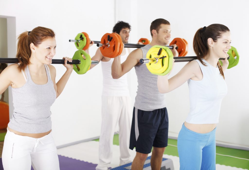 Group of people lifting barbells in weight training class with female trainer in front.