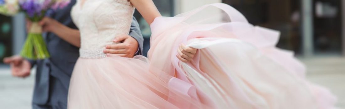 Luxury bride holding a flying pink wedding dress and walking. Beautiful bride in a luxurious dress. Groom hugs bride walking on the street.